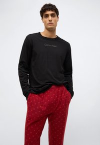 Melna garās piedurknes krekls ar "Calvin Klein" logotipu; sarkani vakara bikses ar atkārtotu mazu "CK" logotipu ornamentu. Mīksta, ikdienišķa auduma.