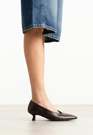Jambes inférieures portant des chaussures à petit talon marron foncé associées à une jupe en jean bleu clair longueur genou sur un sol clair.