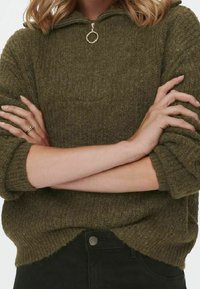 Olivgrüner, gestrickter Pullover mit Halbreißverschluss-Kragen, geripptem Muster und überschnittenen Schultern. Kombiniert mit schwarzen Hosen, die seitliche Schlitze zeigen.