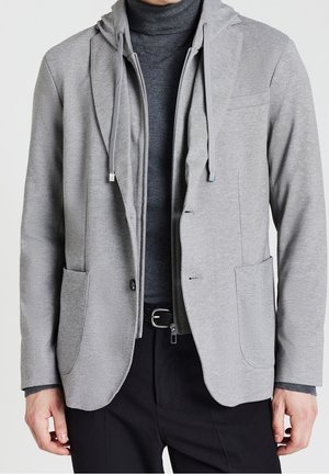 Homme portant un blazer gris superposé avec une capuche attachée, un col roulé foncé, un pantalon noir et une ceinture, debout avec les bras détendus le long du corps.