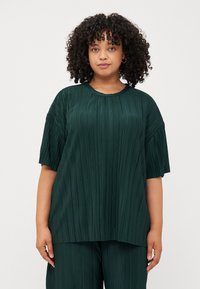 Ένα νέο άτομο με σγουρά μαλλιά φορά ένα πράσινο πλισέ υπερμεγέθες τοπ, ποζάροντας μπροστά από ένα απλό άσπρο φόντο.