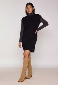 Abito nero senza maniche in maglia, indossato sopra un dolcevita grigio, abbinato a stivali beige in camoscio al ginocchio con tacco a blocco.