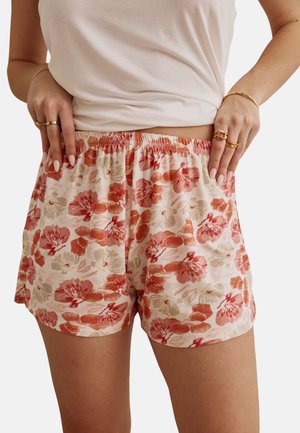 Personne portant un short à motif floral et un haut blanc sans manches, les mains posées sur la taille, montrant des bagues et bracelets en or.