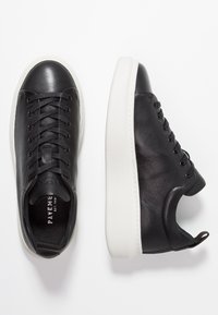 Baskets en cuir noir avec bouts arrondis, semelles en caoutchouc blanches, lacets plats et logo en relief sur la languette, présentant une texture lisse.