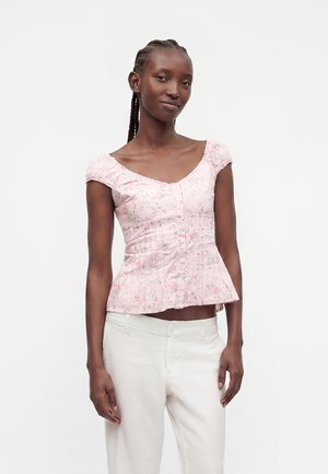 Donna con capelli intrecciati che indossa una camicetta aderente floreale rosa chiaro e pantaloni bianchi, in piedi con espressione neutra su uno sfondo semplice.