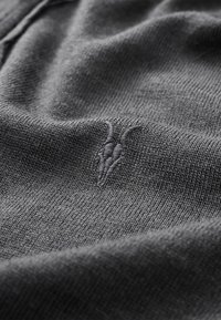 Mörkgrått stickat tyg med en texturerad, broderad design som liknar ett goats huvud. Materialet är mjukt med en slät finish.