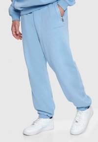 Hellblaue Sweatpants mit lockerem Schnitt, ausgestattet mit einer Reißverschlusstasche und ribbelten Bündchen, kombiniert mit weißen Sneakers.