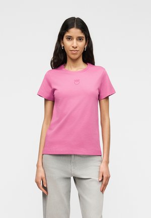 Hosszú, sötét hajú nő egyszínű, világosrózsaszín rövid ujjú pólóban és világosszürke nadrágban, egyszínű világos háttér előtt állva.