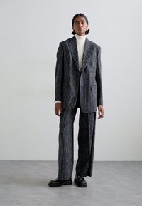 Ραμμένο σκούρο κοστούμι με ρίγες, συνδυασμένο με υπερμεγέθη σακάκι και κομμένα παντελόνια, κατασκευασμένο από υφασμάτινη υφή, σε συνδυασμό με λευκό τοπ με ψηλό λαιμό και μαύρα ογκώδη παπούτσια.