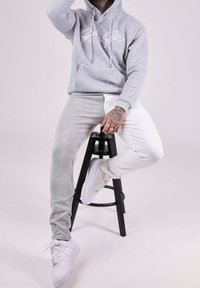 Sudadera gris con logo en script blanco, combinada con jeans: una pierna en gris claro y la otra en blanco. Zapatillas blancas completan el atuendo. Taburete negro.
