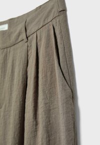 Gros plan sur un pantalon en lin marron à taille haute avec plis et poche latérale sur fond clair.