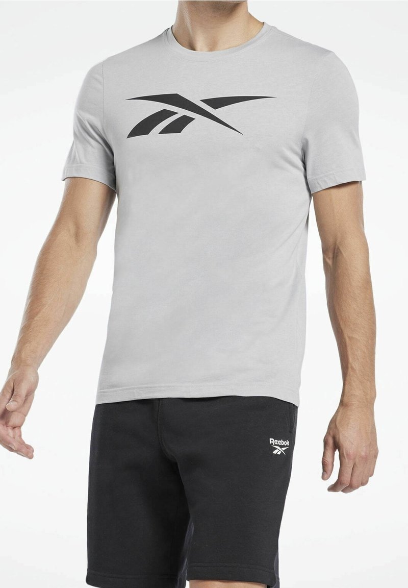 T-shirt in cotone grigio chiaro con logo Reebok nero sul petto, abbinata a pantaloncini da ginnastica neri con logo bianco.