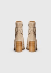 Stivaletti alla caviglia in suede beige con punta rotonda, tacco medio impilato e design con cinturino incrociato che presenta un dettaglio con fibbia dorata sul lato.