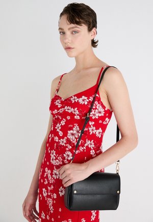 Mujer con un vestido rojo con estampado floral sostiene un bolso de cuero negro cruzado con herrajes dorados sobre su hombro frente a un fondo liso.