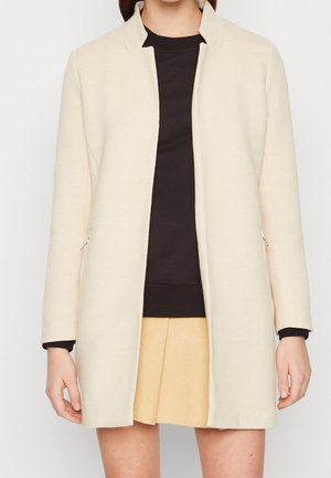 Femme portant un manteau crème ouvert jusqu'aux genoux par-dessus un haut noir et une jupe courte beige, debout devant un fond uni.