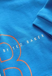 Sudadera de algodón azul con un gran diseño de la letra "B" en naranja y el texto en blanco "BY TED BAKER" impreso en la parte delantera, con una textura suave.