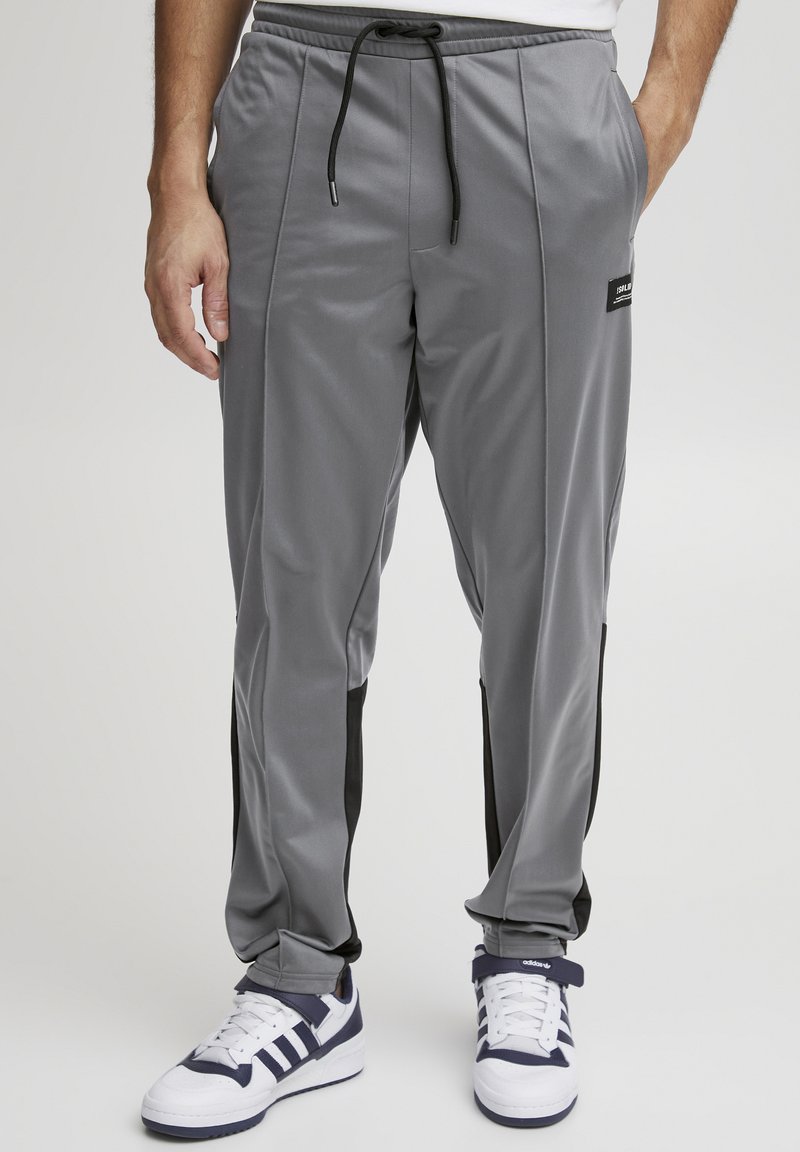 Pantalones deportivos grises con acentos laterales negros, cinturilla elástica, cordón ajustable y bolsillos laterales. Combinados con zapatillas deportivas blancas y marinas.