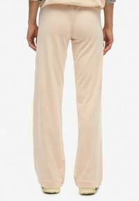 Achteraanzicht van een persoon in losse beige broek en crèmekleurige schoenen, staande tegen een vlakke witte achtergrond.