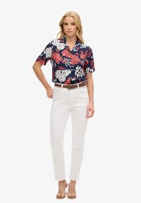 Mujer de pie, vestida con una camisa azul marino con estampado de hojas tropicales rojas y blancas, pantalones blancos de cintura alta, cinturón marrón y tacones con tiras transparentes.