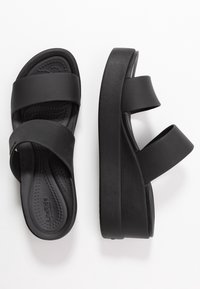 Sandales à plateforme noires à enfiler, avec deux larges brides et semelles moulées, vues de dessus et de côté sur fond blanc.