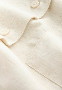 Világos bézs szövet, textúrált szövésű, krémszínű gombokkal és tűzött zsebbel. Puha, sima felület.