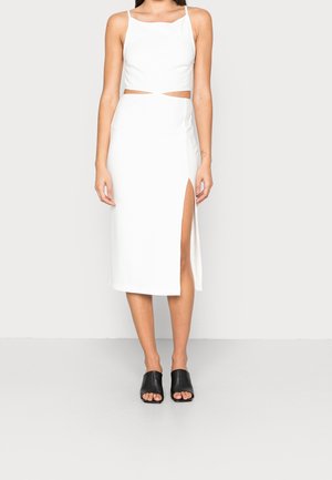 Femme portant une robe midi blanche sans manches avec une découpe latérale et une fente sur la cuisse, associée à des mules noires ouvertes à l'avant, debout devant un fond uni.