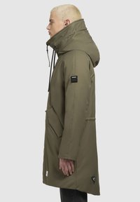Oliven grøn parka-jakke med høj krave, store sidelommer og sorte detaljer. Har en snoretræk og et firkantet logo-patch på ærmet.