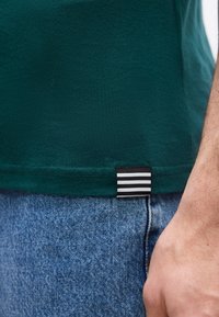 Κοντινό πλάνο ενός ατόμου που φοράει πράσινο πουκάμισο με ετικέτα σε μαύρο και άσπρο ριγέ και μπλε τζιν τζιν, με το χέρι του να ακουμπά στο πλάι.