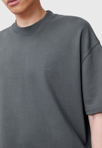 Mörkgrå bomulls t-shirt med rund hals, korta ärmar och präglad logotypdetalj på vänster sida. Slät struktur och avslappnad passform.