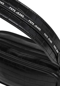 Schwarze Handtasche mit Krokodilprägung, strukturiertem Finish und weißem "Pepe Jeans"-Logo auf dem Tragegurt. Verfügt über ein Reißverschlussfach.