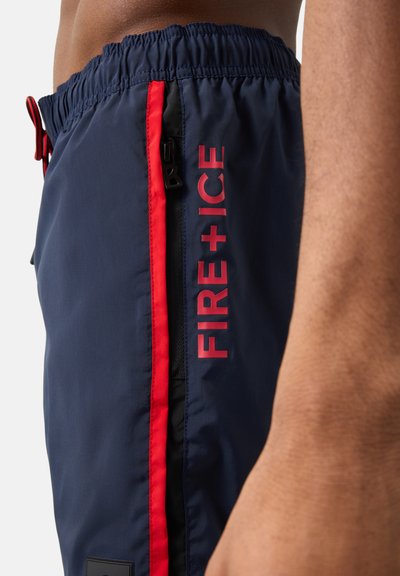 Gros plan sur un short bleu marine avec bordures rouges et texte « FIRE + ICE », doté d'une taille élastique et d'une poche zippée latérale.