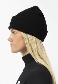 Black knitted beanie with a fold-over cuff, featuring a small white logo patch. Textured fabric and pointed top design.