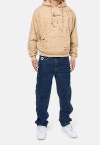 Beige hoodie med ett sprutmönster och framficka, matchad med löst sittande mörkblå denimbyxor och vita sneakers.