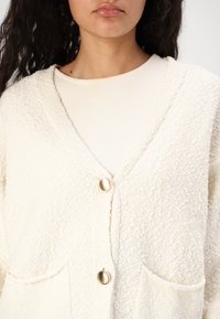 Kremfarvet, textured cardigan med to forlommer, med V-udskæring og guldtonede knaplukninger. Blødt, fluffy materiale.