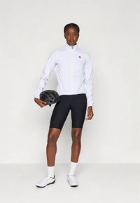 Täiskasvanud naine valges pika käistega jalgrattajacketis ja mustades jalgrattashortsides hoiab musta jalgrattakiivrit, seistes tasase valge tausta ees.