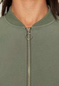 Grön zip-up jacka med ribbad krage, med en silverfärgad dragkedja fram. Slät textur, åtsittande design och inga synliga mönster.