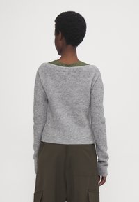 Osoba s krátkými kudrnatými vlasy, oblečená v šedém svetr s dlouhým rukávem a olivově zelených kalhotách, která stojí zády k prostému bílému pozadí.