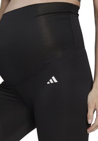 Czarne legginsy ciążowe z wysokim stanem, o gładkiej teksturze, wzmocnionych szwach i białym logo Adidasa po lewej stronie.