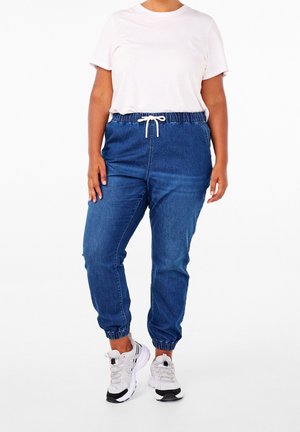 Personne portant un t-shirt blanc, un pantalon de jogging en denim bleu avec taille et poignets élastiques, et des baskets blanches, se tenant devant un fond blanc.