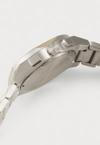Stainless steel wristwatch viewed from the side showing the back cover, two rectangular buttons, and a round crown on the case.