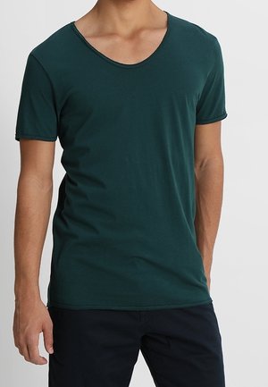 Mężczyzna w ciemnozielonej koszulce z krótkim rękawem i dekoltem w serek oraz czarnych spodniach, stojący z jedną częściowo widoczną ręką.