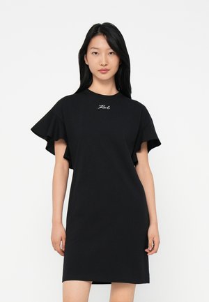 Young woman with long black hair wears a black short dress with ruffled sleeves and small white embroidered text on chest.
