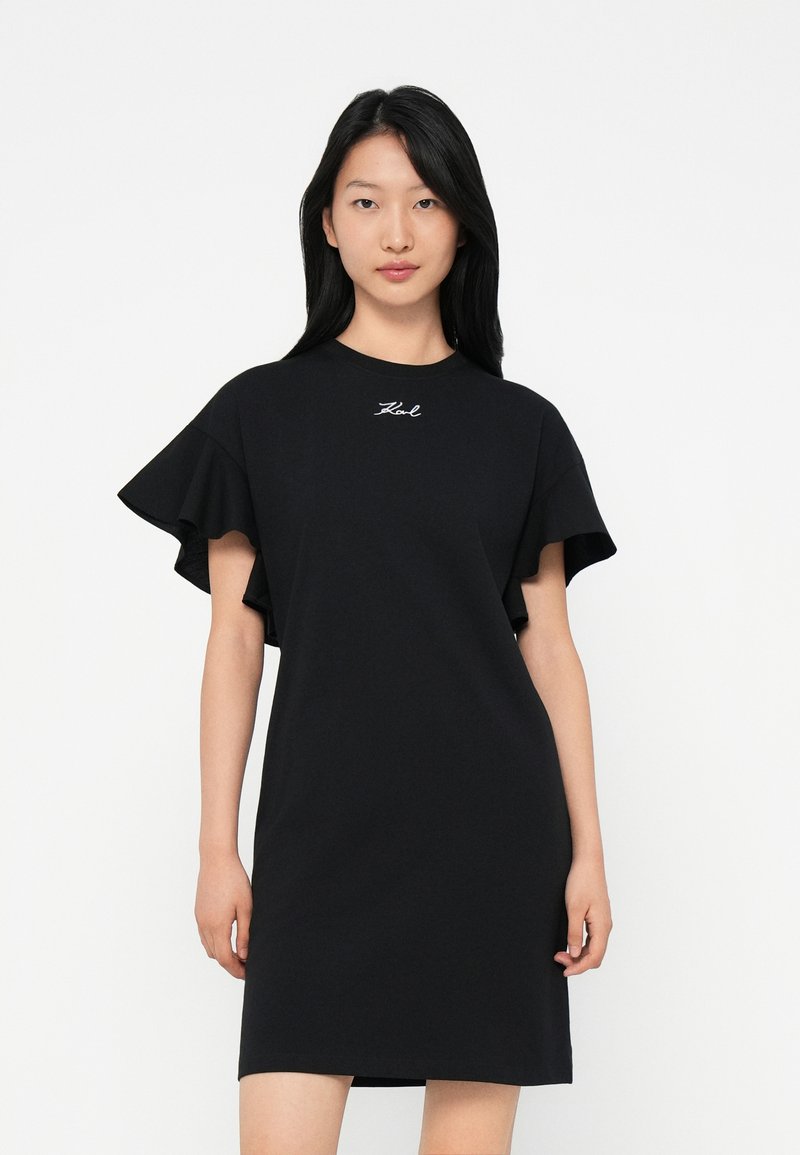 Νεαρή γυναίκα με μακριά μαύρα μαλλιά φοράει μια μαύρη κοντή φόρμα με ρυτιδωμένα μανίκια και μικρό λευκό κεντημένο κείμενο στο στήθος.