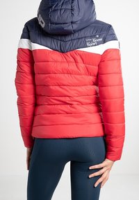 Polstret rød vinterjakke med hætte i marineblå, hvide detaljer og logo. Har en quiltet tekstur og en figursyet pasform.