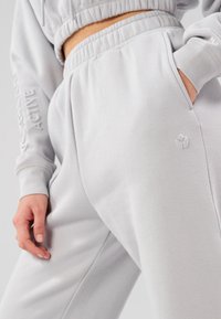 Pantalon de survêtement gris clair en tissu doux, avec une coupe décontractée, une taille élastique et des poches latérales. Détail discret du logo de la marque visible.