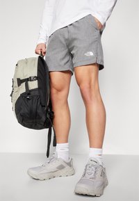 Pilkos šortai su The North Face logotipu, derinami su pilkais sportbačiais ir baltais kulkšninių kojinių poromis. Juoda ir alyvuogių žalio atspalvio kuprinė su mikro tinklelio akcentais.