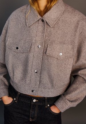 Persoon draagt een cropped, grijs wollen jack met drukknoopsluiting en klepzakken, gecombineerd met zwarte denim jeans, handen in de zakken.