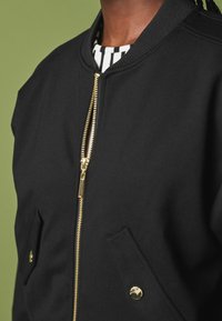 Chaqueta bomber negra con una cremallera dorada, cuello acanalado y dos bolsillos frontales. Presenta una textura suave con un diseño minimalista.