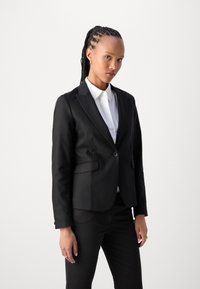 Černý ušitý blazer z hladké látky, s jednobarevným zapínáním, klopami a dvěma předními kapsami. Nosit s bílou košili.