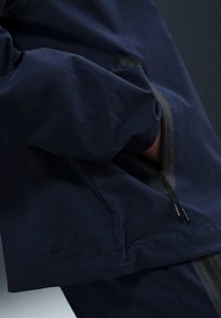 Marinblå jacka med en jämn textur, utrustad med en dragkedjeficka och en diskret logotyp. Designad med kontrasterande grå detaljer.